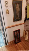 Photo showing two framed brass rubbings, one hung on a white wall and one leaning against the wall on a hardwood floor.