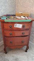 Front view of a vintage wooden side table with three drawers, showing metal ring pulls and a green protective mat on the top surface covered with jewelry items.