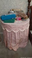 Front view of round plywood side table covered with pink scalloped and floral patterned fabric cover, various items on top not included