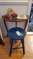 Wood demi-lune side table with curved legs, a lower shelf, and various small decorative items on top. Painted blue wood stool in front with round seat and three legs.