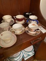 View of seven vintage porcelain teacups with matching saucers. Varied floral and gold decorative patterns. Cups arranged on wooden shelf.