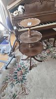 Full view of the three-tiered wooden pie crust table standing on a floral carpet in front of a piano.