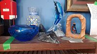 Shelf displaying the blue glass bubble ashtray, glass dolphin figurine on a glass base, dark carved soapstone figurine, and glass vase with diamond pattern and yellow accents.