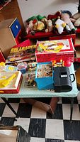 Overview of vintage toys and games laid on table including boxed Mattel Big Loader construction set, Preschool Camp Hot Pot game, assorted puzzles, cash register toy, and several plush dolls on red bench in background.