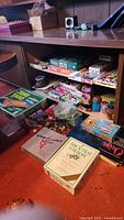 Photo showing various vintage boxed board games and toys stacked under a shelf, including Junk Yard, Perfection, Clue, and Tiddly Winks.