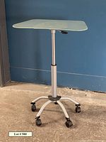Adjustable height workstation table shown near wall, glass top at raised height, caster wheels visible on base