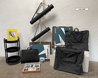 Full lot view showing black round 3-tiered metal shelf, two black cylindrical canvas carrying tubes, several black portfolios, a mat cutter, drawing paper pad, and tray with pencils and tools on concrete floor.