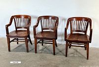 Front view of three vintage solid wood banker’s chairs attached side by side with visible slatted backs, flat wooden seats, and armrests on at least one chair.
