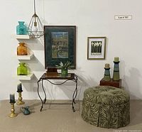 Room scene showing three Blenko Glass pitchers on white floating shelves in teal, amber, and lime green, framed prints on wall, iron leg table underneath with decorative boxes and faux plant, green leafy fabric storage ottoman on floor, gold candlesticks and ceramic striped candlesticks visible.