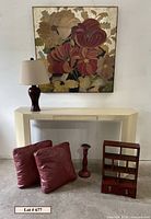 View of blonde lacquered console table with one drawer, underneath two maroon pillows, maroon candlestick, small Asian cubby shelf. Above table large floral artwork, on table lamp with maroon base and off-white shade.