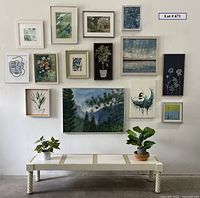 Full view of the gallery wall with 14 framed artworks arranged above a low plant table holding two faux plants