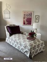 Chaise lounge with floral fabric, purple velvet pillow, small floral arrangement on chaise. Framed Boheme artwork and smaller framed watercolor on the wall, white wire birdcage on wall, white lamp with shade on white side table.