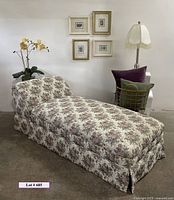 Front-left angle of floral patterned armless chaise lounge with accessories in the background against white wall.