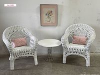 Two white wicker children’s chairs with pink lumbar pillows flanking a small white table, with framed Picasso-style floral print above.