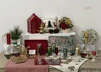 Wide shot of the Christmas decor collection showing the red house lantern, snowman pillow, brass snowman teapot set, faux floral arrangement, champagne flutes, candles, and assorted linens and trays laid out on white tiered tables.