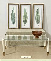 View of coffee table under triptych artwork with wooden pedestal bowl on table, showing glass top, metal frame, and gold-tipped legs.
