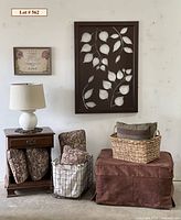 Photo showing nightstand with lamp, wire basket with pillows, wicker basket on ottoman, small wall art, large laser-cut metal leaf decor on wall.
