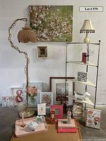 Overall photo of lot showing various framed artwork, metal corner shelf, metal floor lamp with wicker shade, small ceramic lamp with beaded shade, and decorative items displayed on and around the shelf