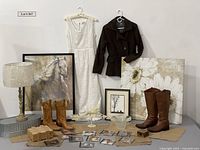 Full display of lot items including clothing dress and jacket, boots, art pieces, lamp, and smaller household items arranged on table and hung on wall.
