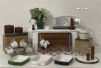 Overall shot of entire lot showing wooden trays, enamel casserole pans, glass tumblers, ceramic coffee set, glass pitcher, wooden canisters, salt and pepper shakers, microwave pot, teapot, creamer, faux plants, candles, and miscellaneous decorative items.