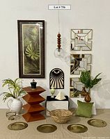 Full lot showing Eye of Horos wooden wall decor, sculptural occasional table, brass chargers, pendant light with glass globe, framed leaf art print, pitcher, foliage, mirrors, pyramid, urn, basket, pillow, and decorative container.