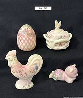 Four pieces of Fenton Burmese art glass on black background: an egg-shaped piece with floral motif, a butterfly floral covered dish, a rooster figurine, and a bear figurine lying down.