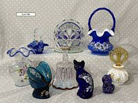 Overview of all Fenton glass items including vases, baskets, perfume bottle, animal figurines, bell and fleur de lis display on dotted background