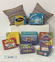 Eight boxed board games arranged with two colorful feather pillows behind them, well-lit showing front of boxes and pillow patterns.