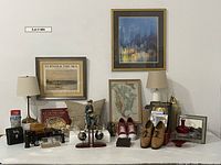 Full view of lot items including framed artworks, lamps, glassware, shoes, binoculars, books, and decorative objects on a table against a white wall.