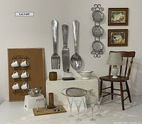 Photo showing full lot including large silverware wall decor (fork, knife, spoon), wooden chair, crystal glasses, bulletin board with mugs, framed artworks, and assorted kitchen items.