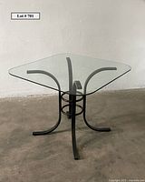 Square glass-top table with rounded edges and black metal base with curved legs and circular support near center, shown on concrete floor against plain wall.