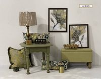 Full display showing green painted wooden table with decorative items on and around it including brass lamp, patterned pillows, framed prints, ottoman, wire basket, and decorative knobs box.