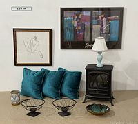 Full view of lot items including VonHaus stove heater, three teal velvet pillows, framed artworks, decorative wire baskets, planter, and lamp.