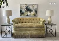 Green velvet vintage sofa with rolled arms, tufted back, fringe skirt, paired with two metal ring design end tables and two creamy white lamps with shades, with floral canvas artwork above. Shows general condition and detail of fabric wear but no rips or tears.