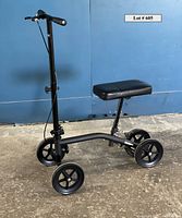 Side view of black knee walker showing four wheels, handlebar, and knee platform against blue wall.