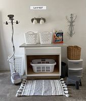 Wide view of laundry folding station showing cabinet with baskets on and in it, garment steamer to left, wicker basket on top of Sterilite drawers, box of Tide cleaner, wall light above and hooks to right, step stool and rug in front.