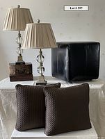 Two lamps, decorative box, leather ottoman, and two pillows arranged together on cloth-covered table.
