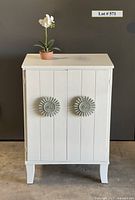 Full front view of solid white wood cabinet with two large decorative medallion handles on the front doors, standing on four short legs.