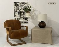 Full view of the lot with the burnt orange cantilever chair, abstract art on wall, hanging planter with faux plant, and side table with lamp