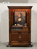 Front view of the wood armoire bar cabinet with framed Mona Lisa print visible inside glass door, hanging wine glasses above, rock glasses lined below, silver trays, and an ice bucket on top shelf.