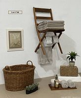 Full view of vintage wooden folding chair with towels, large wicker basket, small woven basket with ceramic planter and faux plant, teak tray with bathroom items, small framed floral artwork, and oval planter with faux succulents.
