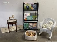 Wide view of entire lot showing all items together including book rack with colorful children's books, white wicker rocking chair with stuffed rabbit, basket of wooden blocks, round marble topped table with wooden rocking horse figurine, and wooden sign.