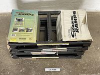 Pair of black folded steel loading ramps with original packaging on top, placed horizontally on floor.