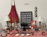 Full display of items including black and white inspirational poster, color block apron, glassware, ceramic mugs, small lamp, decorative items spread on pink cloth.