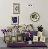 Photo showing multiple purple-themed decorative items including framed floral artworks on the wall, an arrangement of throw pillows with floral and abstract purple designs, faux purple flowers in pots and vases, a candle, small framed drawings, and a decorative box with purple roses on a gray surface.