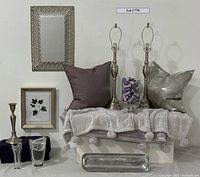 Full lot overview showing all items displayed on and around two white boxes; including crystal vases, pillows, throw blanket, candle holder, metal tray, lamp bases, metal mirror, framed botanical print, and glass vase with threads.