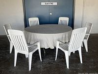 Front view showing large round folding table covered in light-colored linen tablecloth surrounded by six white plastic outdoor chairs.