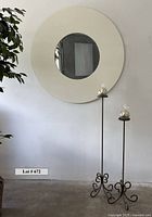 Round wall mirror with creamy white outer frame and smaller mirror center on a plain wall, with two tall iron candlesticks holding new round candles to the right.