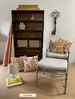 Wooden shelf filled partially with some decor, iron chair with cushions, floral pillows, fabric rolls, lamp and small decor items arranged around.