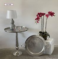 Full view of silver accent table holding pink cocktail glasses and gray lamp, with matching silver tray on floor and faux raspberry orchid in background